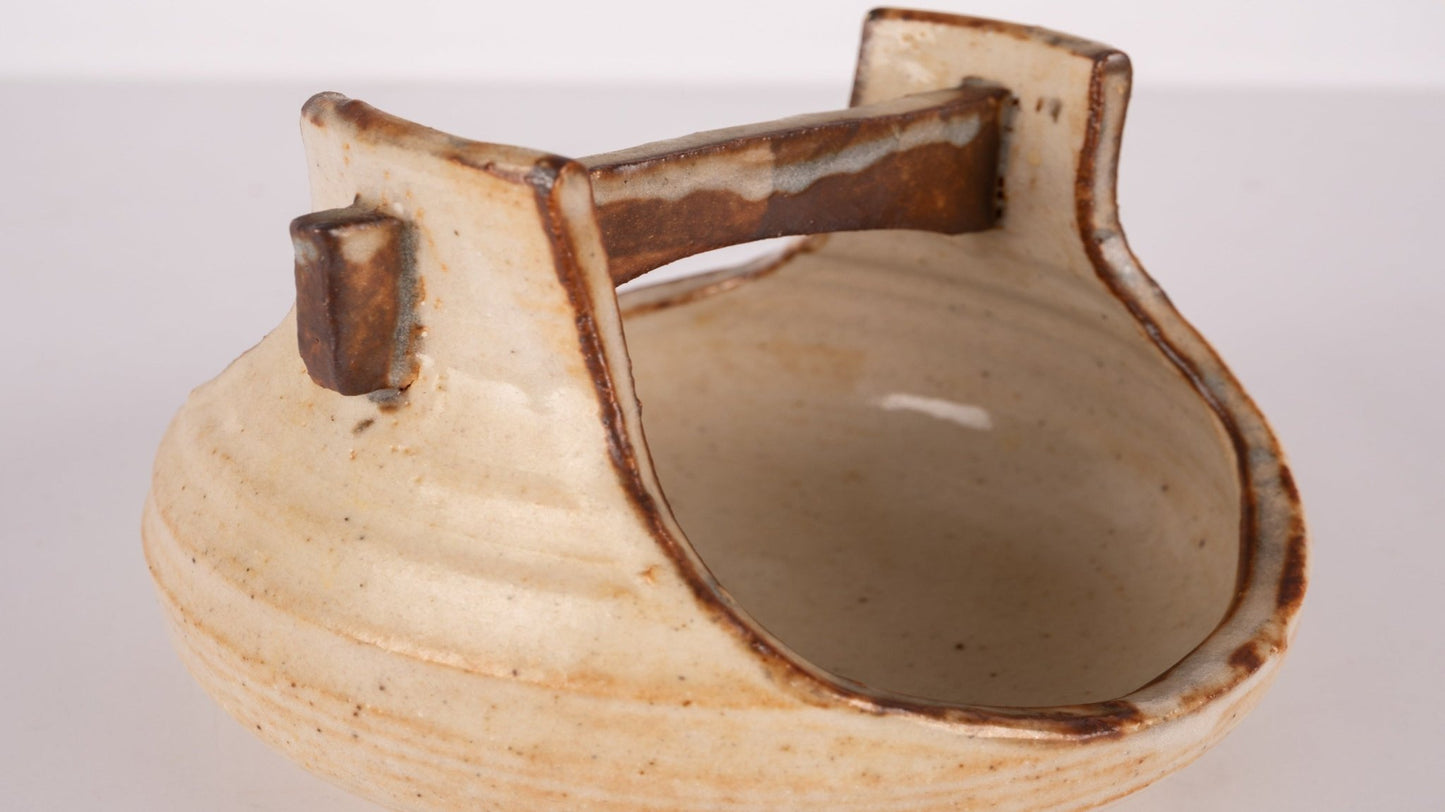 Small studio pottery bowl with handle, natural beige glaze, and rustic brown accents on white background