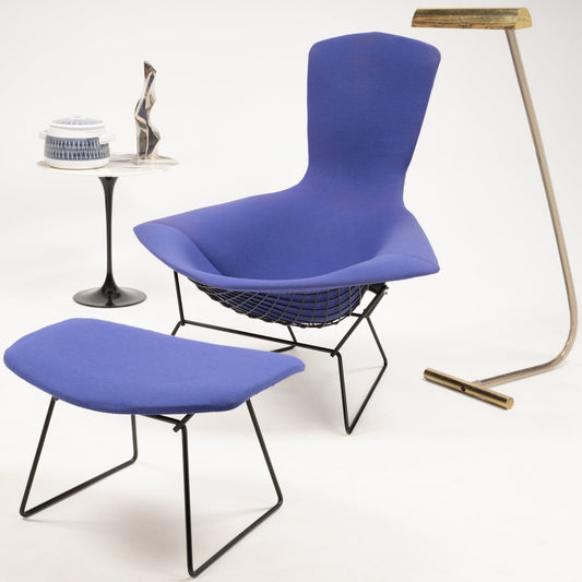 A modern design room featuring a blue lounge chair and matching footrest with black metal frames. Nearby, a black side table with decor and a sleek floor lamp.