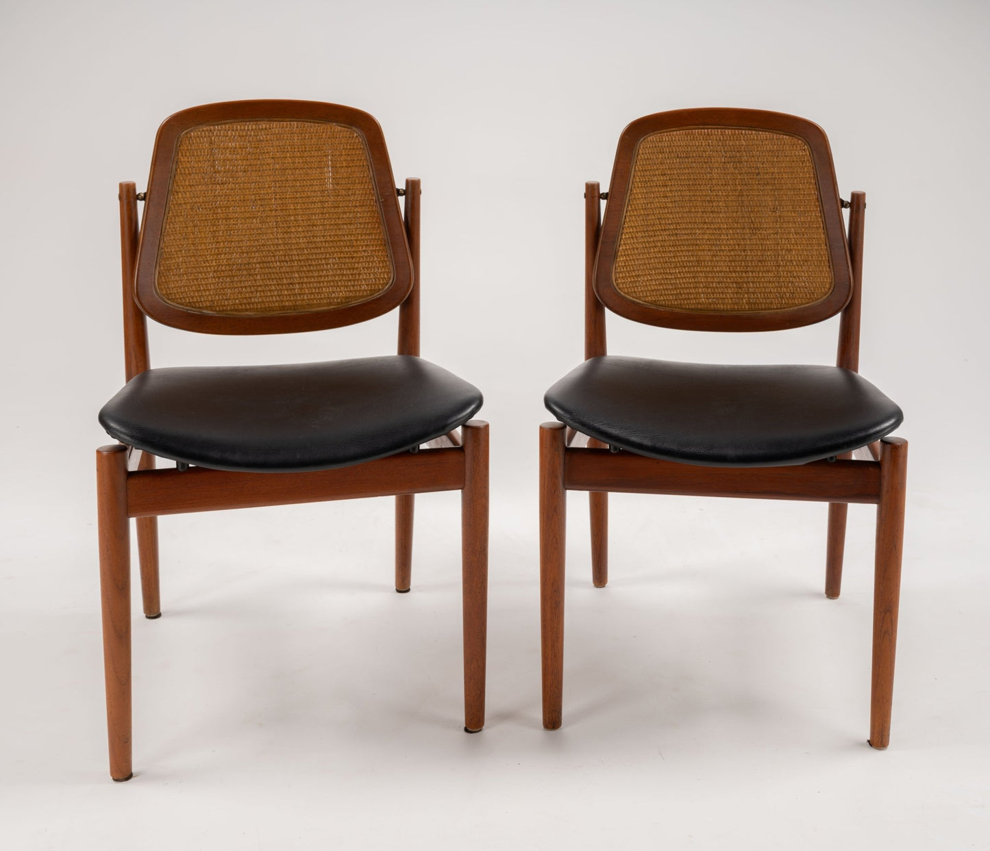 Pair of mid-century modern teak dining chairs with woven cane backrests and black leather seats on white background