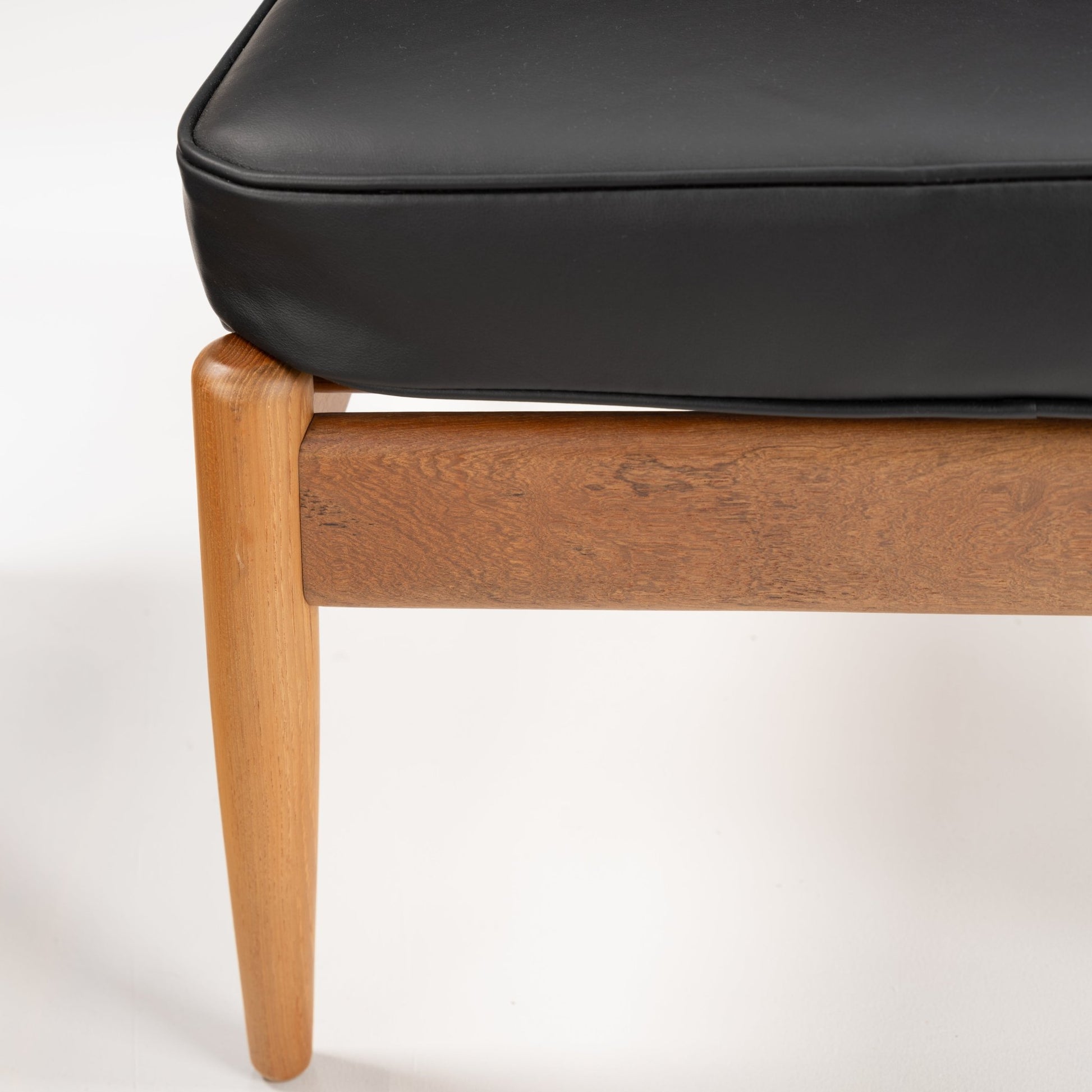 Danish Teak Ottoman with Black Leather featuring a close-up of tapered teak legs supporting a smooth black leather cushion, highlighting vintage mid century modern furniture craftsmanship.