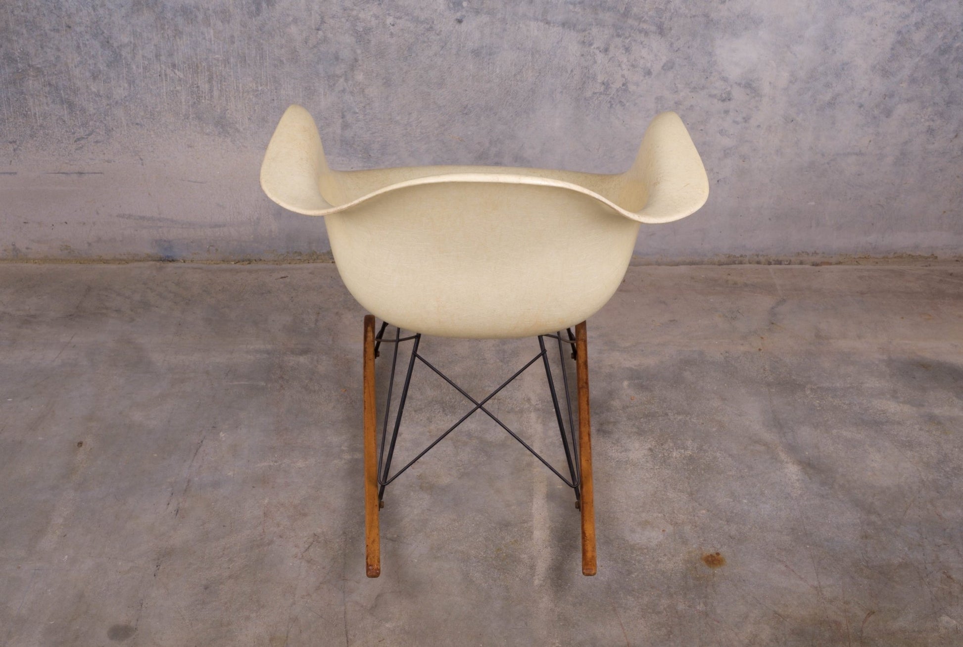 Eames rope-edge fiberglass rocker with wooden base and metal supports on concrete floor against textured wall
