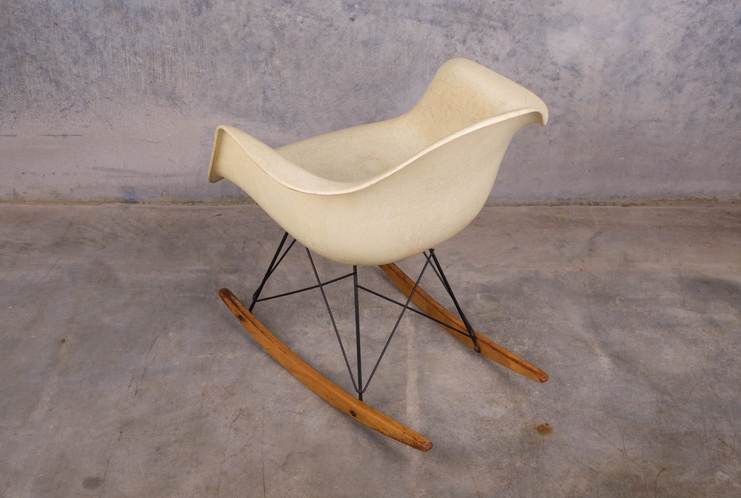 Mid-century modern Eames fiberglass rocking chair with rope edge and wooden rocker base on concrete floor background.