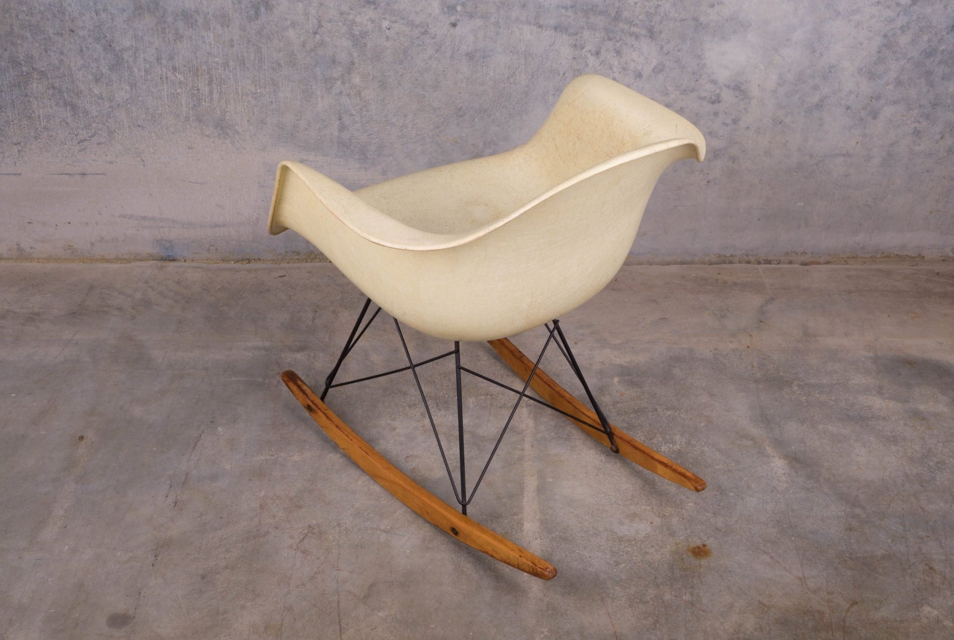 Mid-century modern Eames fiberglass rocking chair with rope edge and wooden rocker base on concrete floor background.