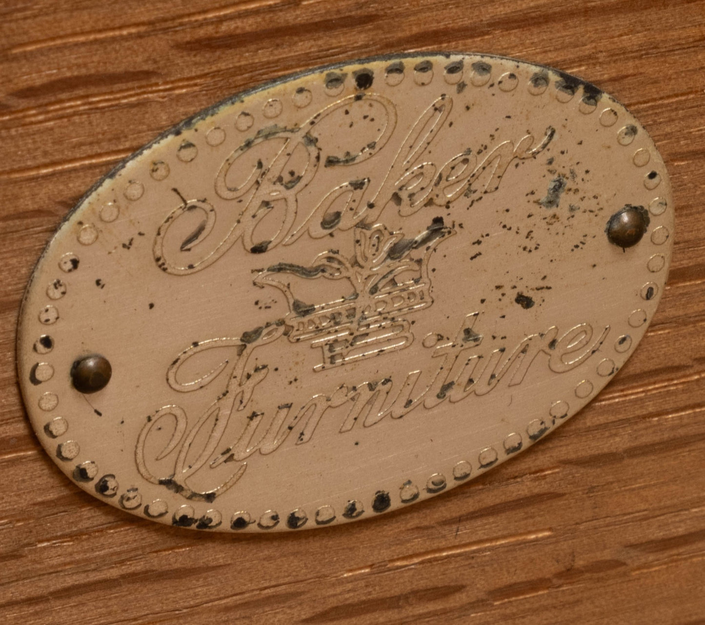 Oval vintage metal Baker Furniture brand plaque with crown emblem mounted on walnut wood surface