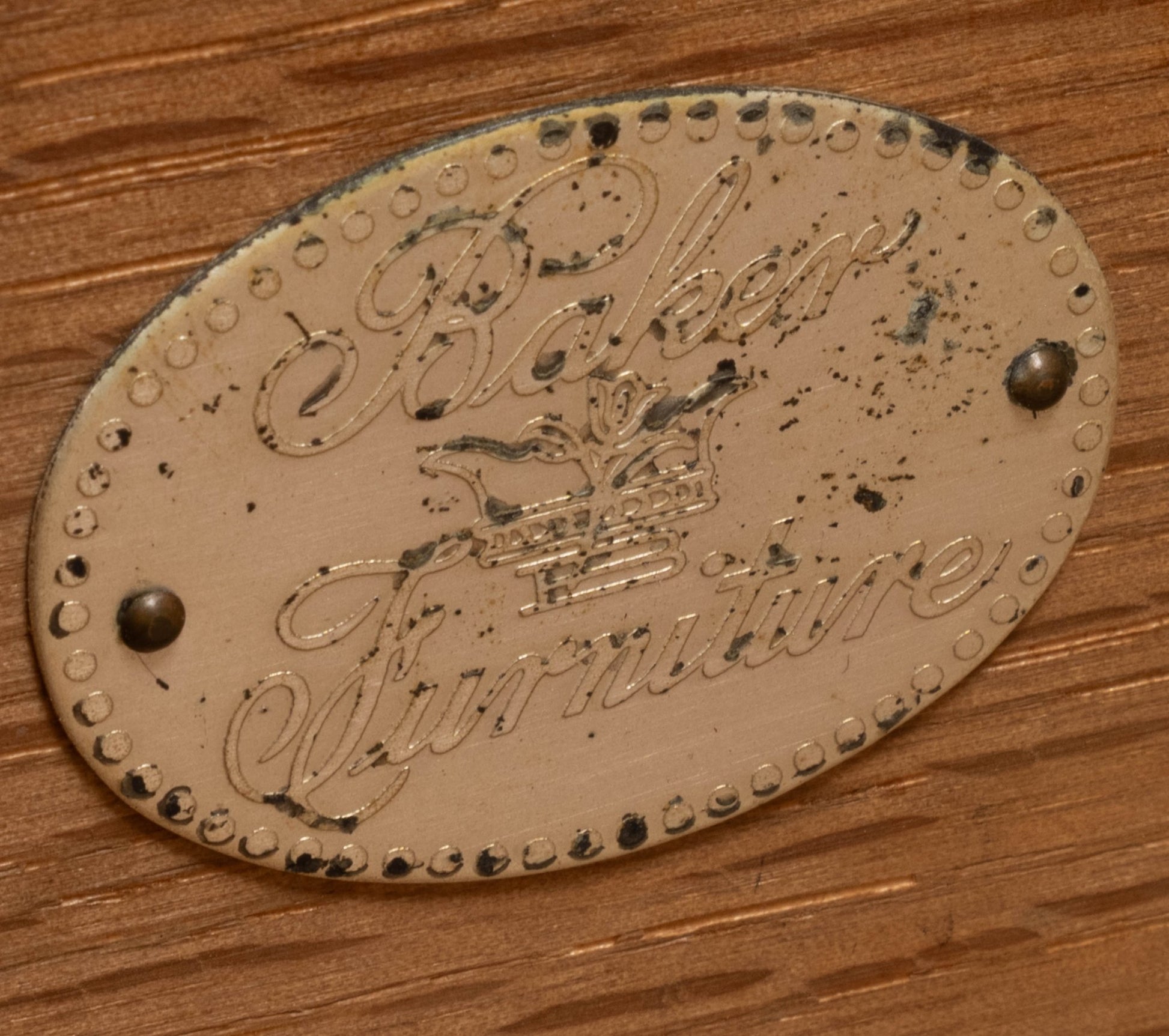 Oval vintage metal Baker Furniture brand plaque with crown emblem mounted on walnut wood surface