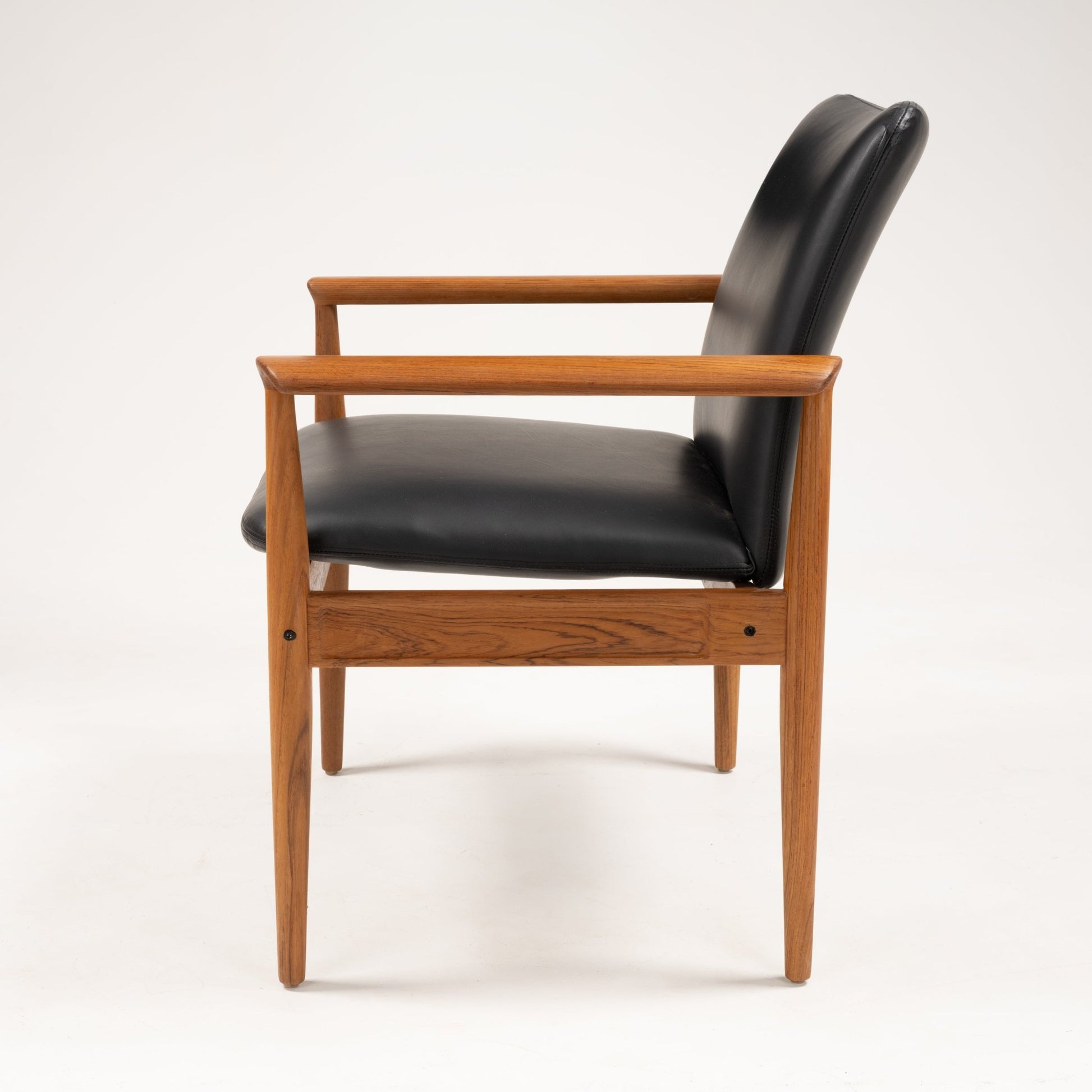 Side profile of Finn Juhl Model 209 "Diplomat" chair with teak frame and black leather seat and backrest on white background