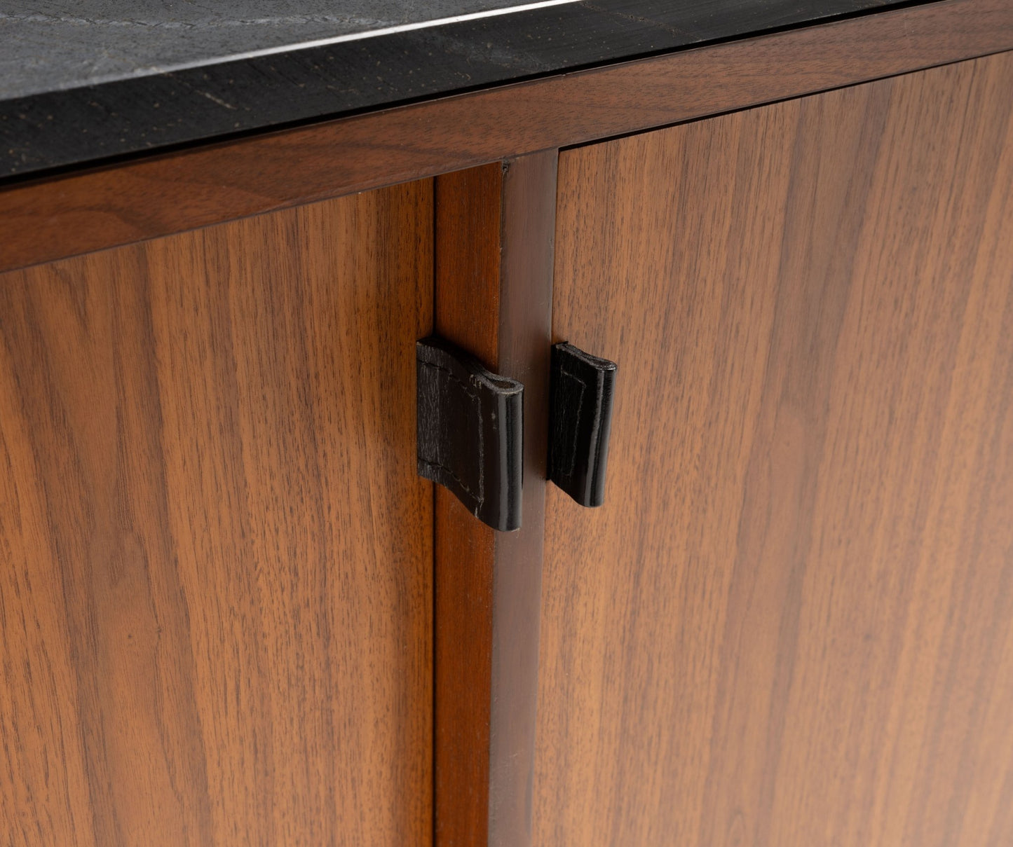 Close-up of mid-century walnut credenza doors with original black leather pulls and black marble top edge visible