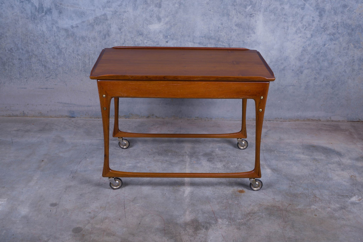Mid-century modern Ingvar Jensen teak bar cart with sliding top, black laminate, and rolling wheels on concrete floor