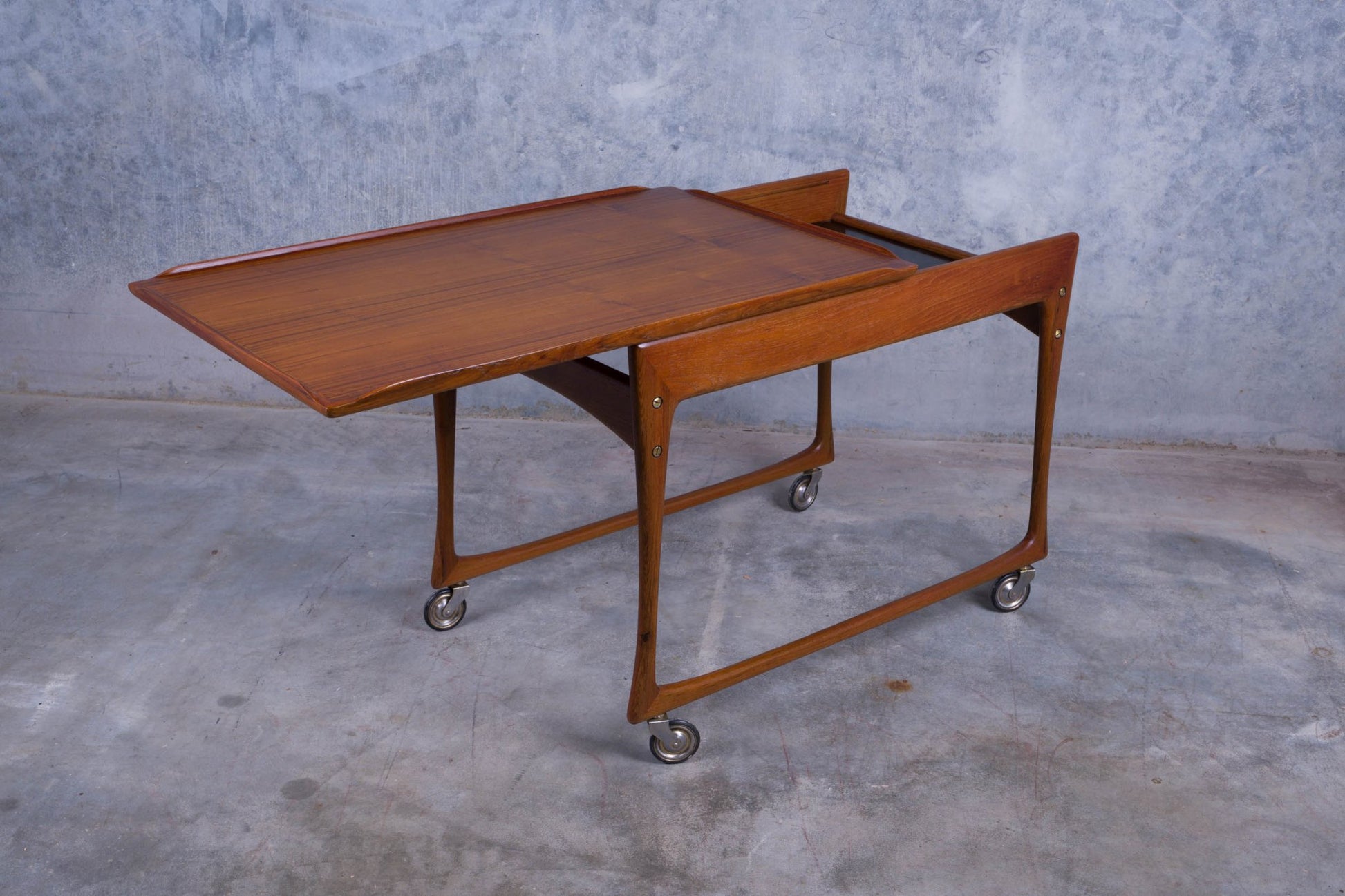 Mid-century modern Ingvar Jensen teak bar cart with sliding top and black laminate compartment on caster wheels.