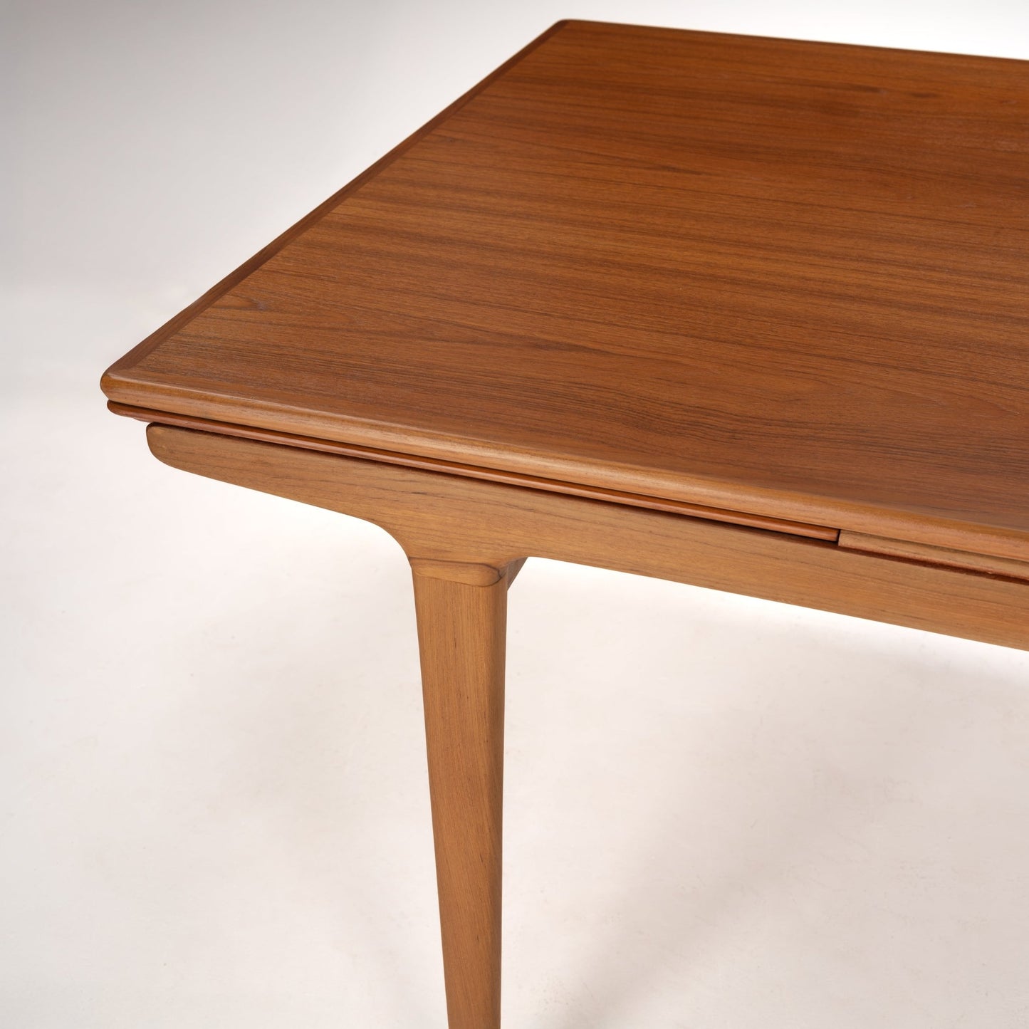 Corner view of smooth wooden table top with subtle beveled edge and slender tapered leg in warm natural finish
