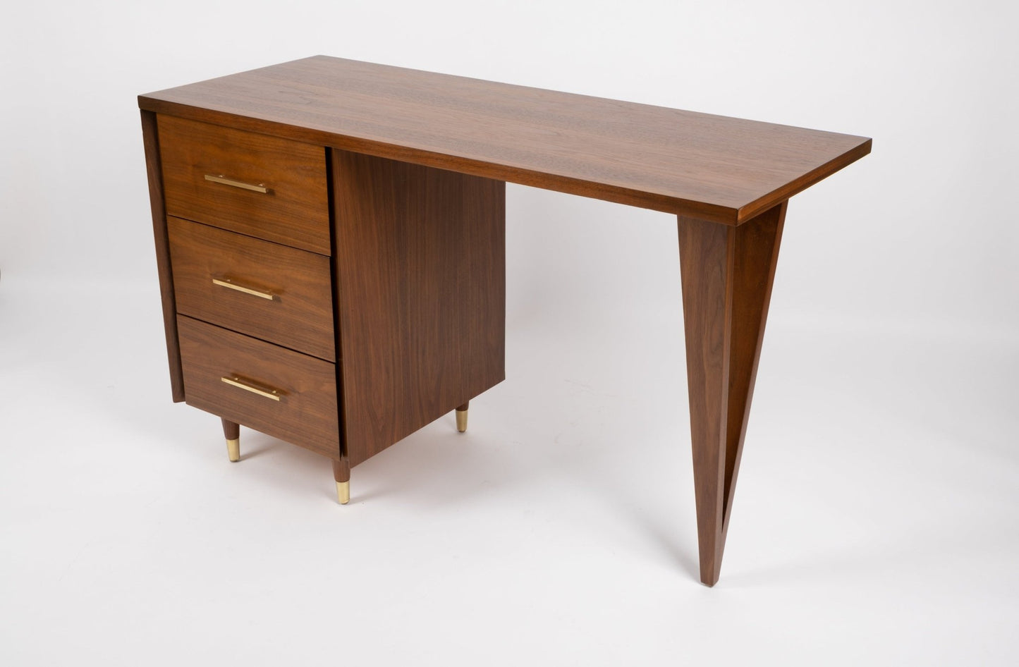 Mid-century modern wooden desk with three brass-handled drawers, tapered triangular leg, and brass feet on white background