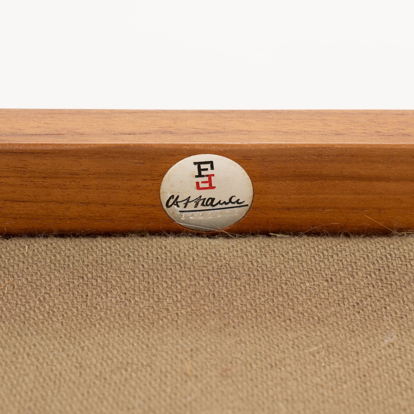 Close-up of a circular metal label with black and red emblem and cursive signature on wooden furniture frame