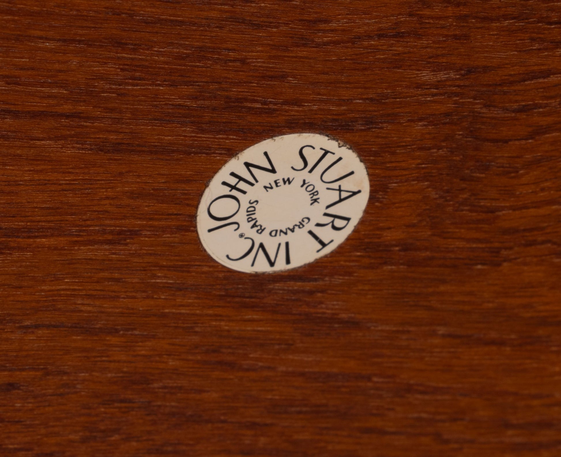 Close-up of John Stuart Inc New York label on solid teak furniture surface with rich wood grain texture