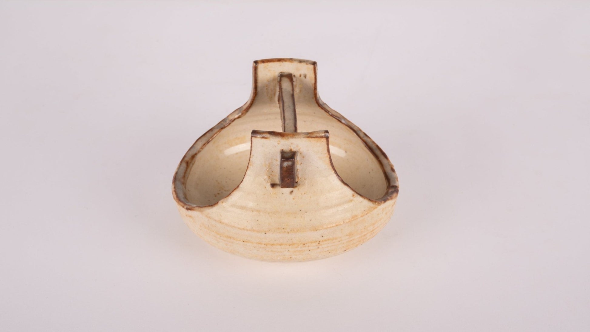 Small studio pottery bowl with handle in cream glaze, featuring stamped signature on the bottom, on plain background.