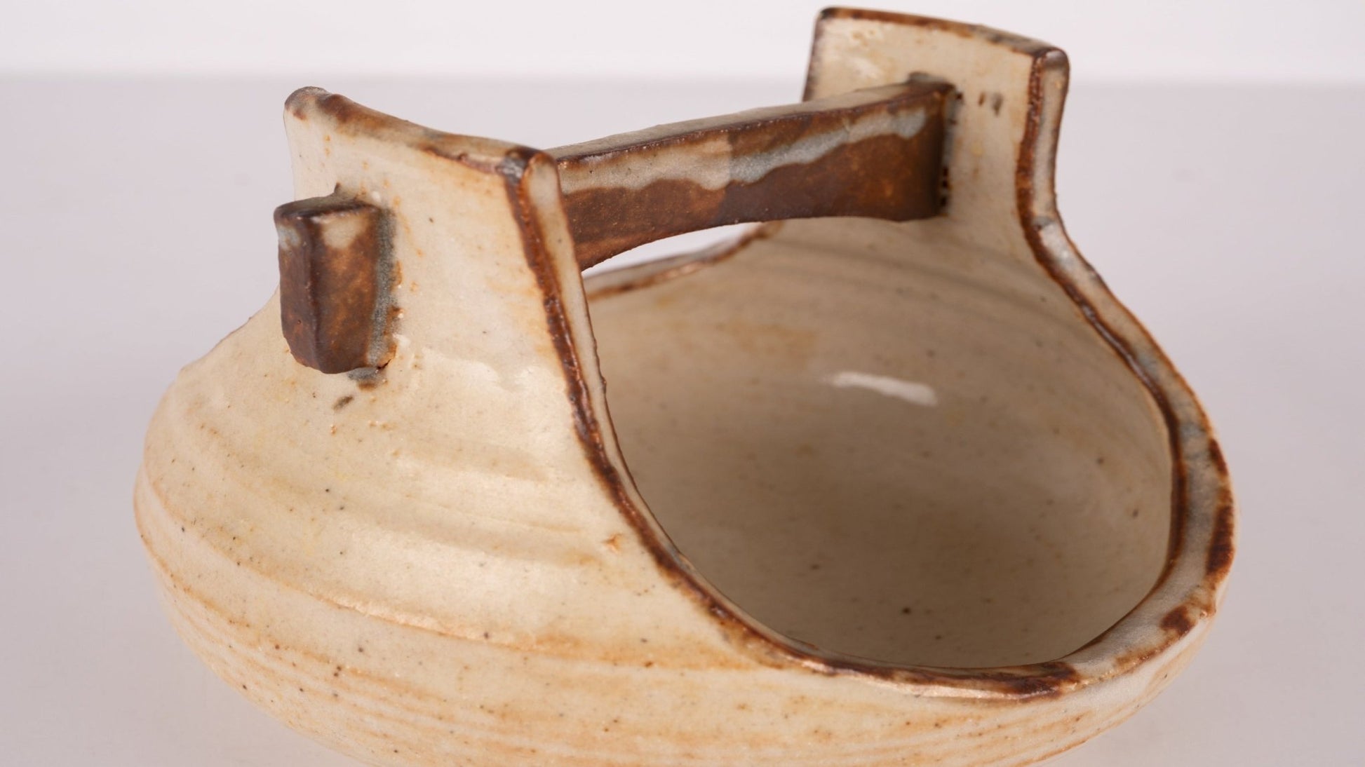 Small studio pottery bowl with handle, natural beige glaze, and rustic brown accents on white background