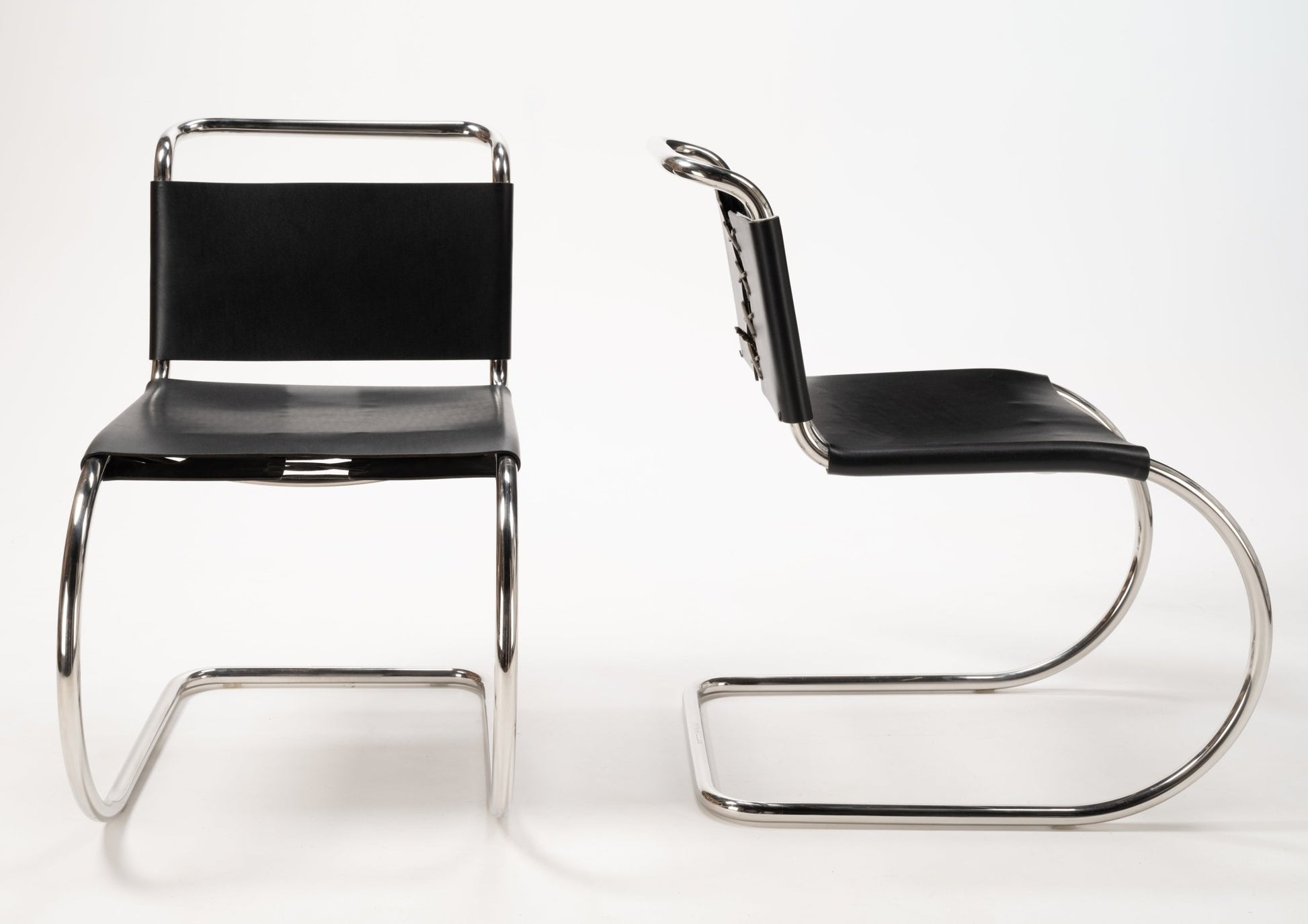 Two Van Der Rohe MR10 cantilever chairs with polished chrome tubular frames and black leather seats and backs