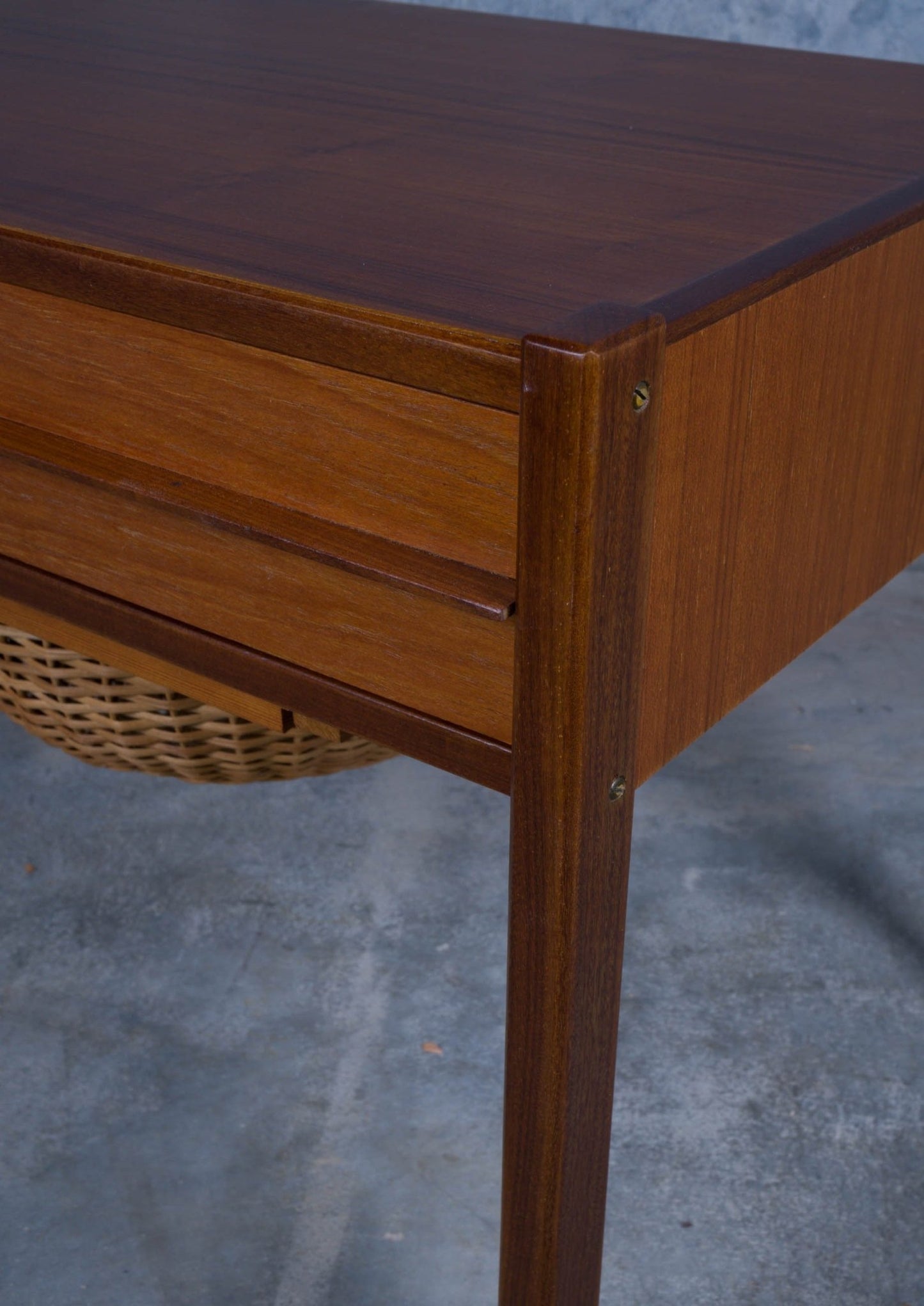 Close-up of Danish teak sewing side table corner showing drawer front and woven cane circular container below.