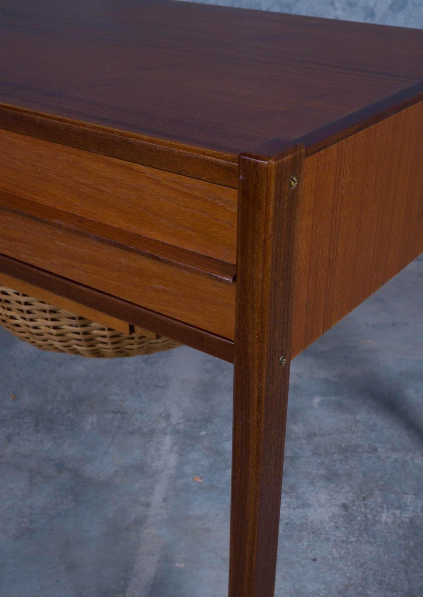 Close-up of Danish teak sewing side table corner showing drawer front and woven cane circular container below.