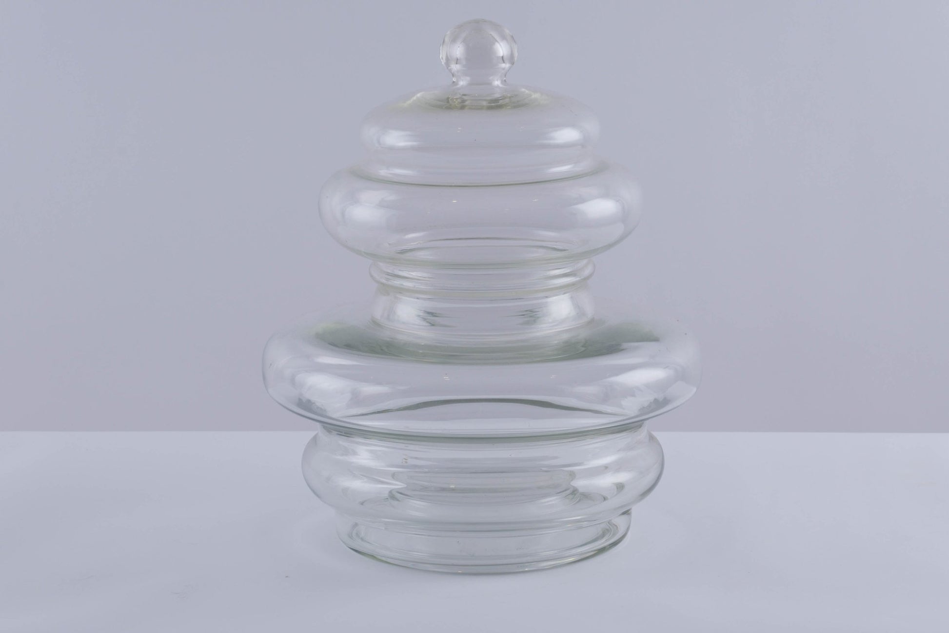 Large clear glass jar vessel with rounded lid and smooth tube-shaped body tapering outward from base.