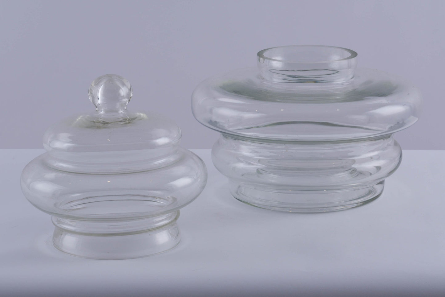 Set of two clear glass jar vessels with rounded, tapered shapes and one featuring a ball handle lid on white surface