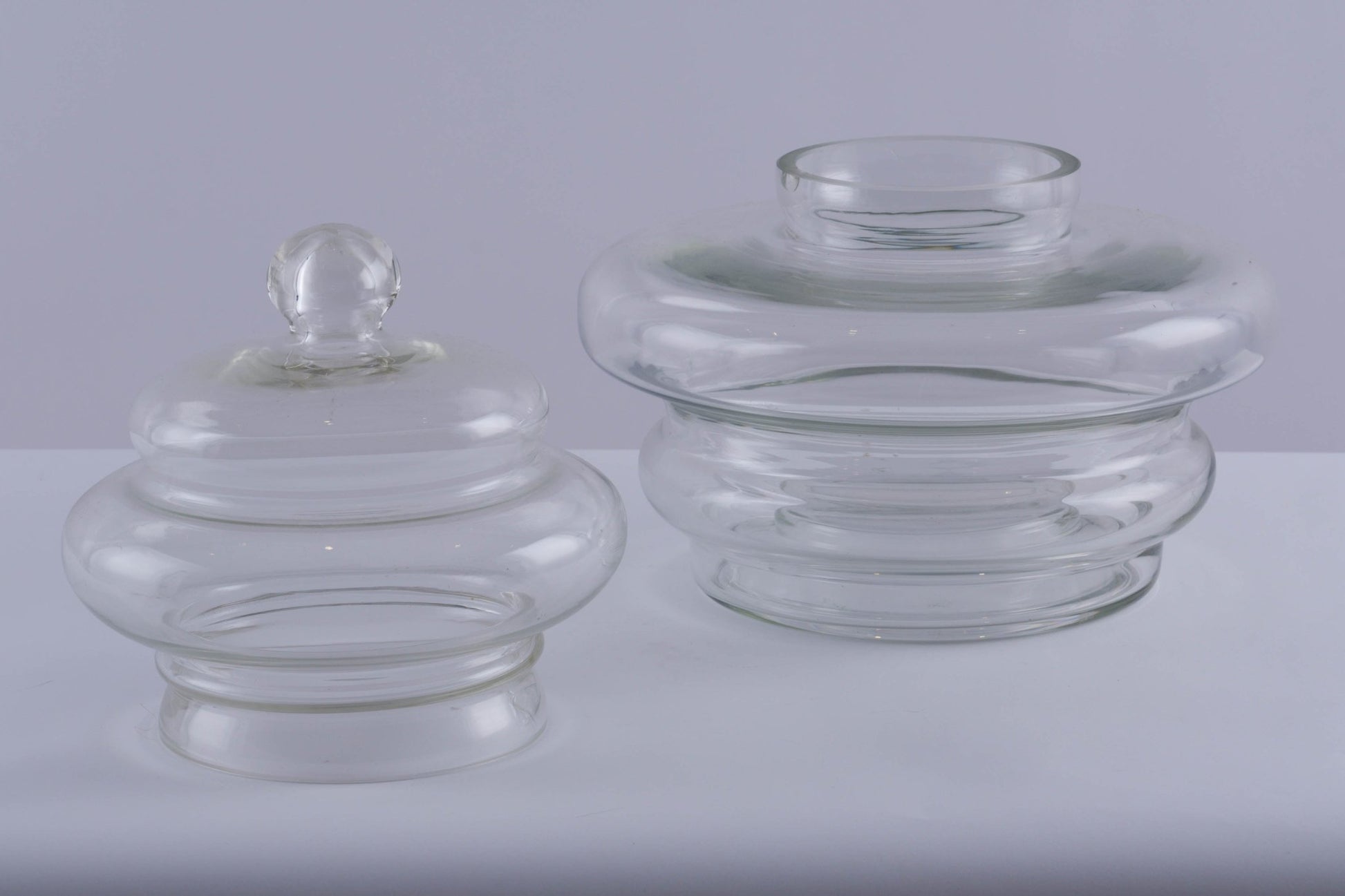 Set of two clear glass jar vessels with rounded, tapered shapes and one featuring a ball handle lid on white surface