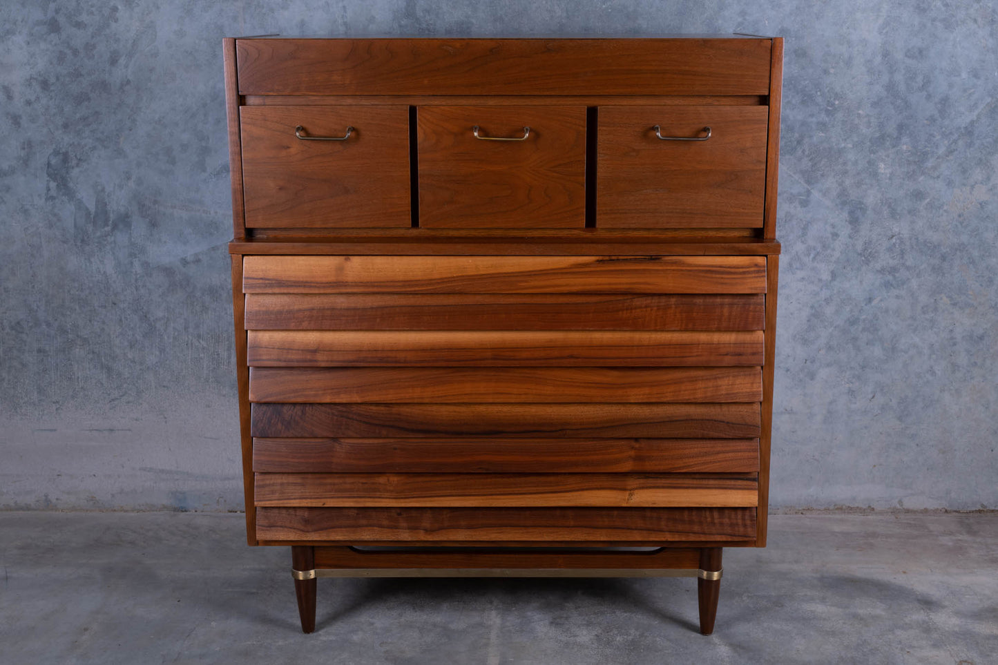 Mid-century modern walnut louvered highboy dresser with seven drawers and brass handles on tapered legs.