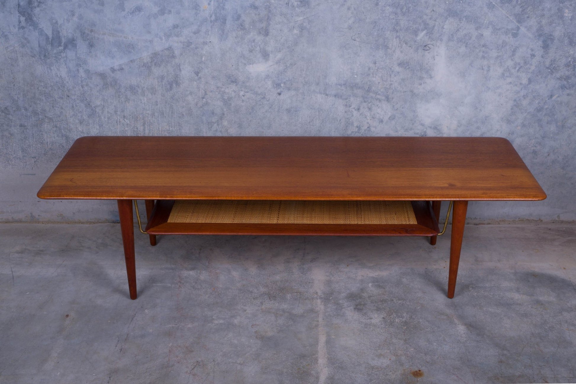 1950s Danish teak coffee table with cane shelf, tapered legs, and minimalist mid-century modern design on concrete floor