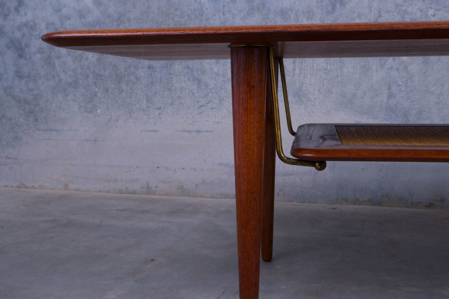 Close-up of mid-century modern teak coffee table leg with brass detail and cane shelf on concrete floor background